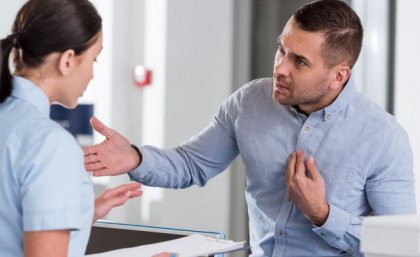 A man looking aggressive and threatening stands before a woman at a medical reception desk.